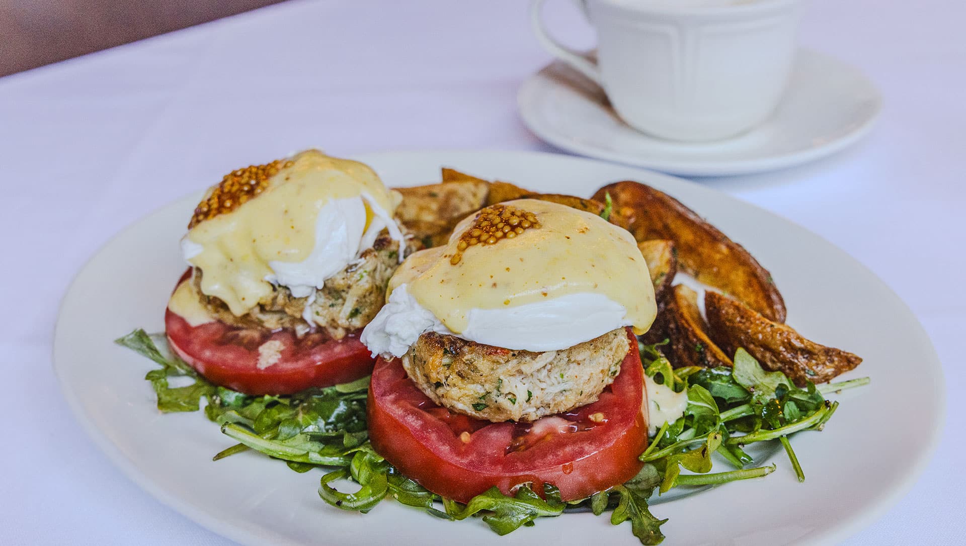 Two eggs Benedict with poached eggs, hollandaise sauce, and mustard seeds on tomato and greens, with potato wedges, served on a white plate. Coffee cup in the background. Two eggs Benedict with poached eggs, hollandaise sauce, and mustard seeds on tomato and greens, with potato wedges, served on a white plate. Coffee cup in the background.