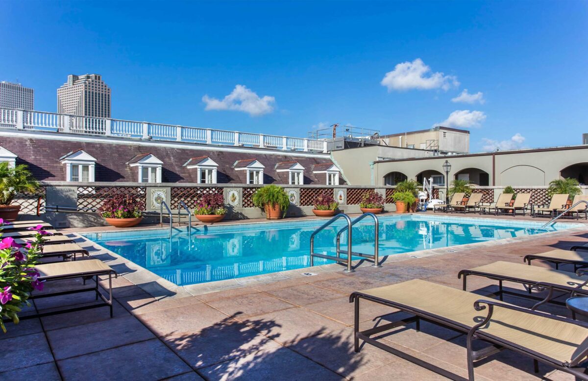 Rooftop swimming pool surrounded by lounge chairs and potted plants, with city buildings visible in the background under a clear blue sky.