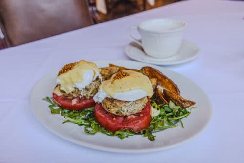 A plate with two eggs Benedict on tomato slices and greens, served with roasted potatoes, and a cup of coffee in the background.