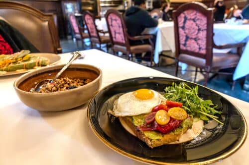 A plate with avocado toast topped with vegetables and a fried egg, served with a side salad, sits next to a bowl of oatmeal on a white tablecloth in a restaurant dining room.