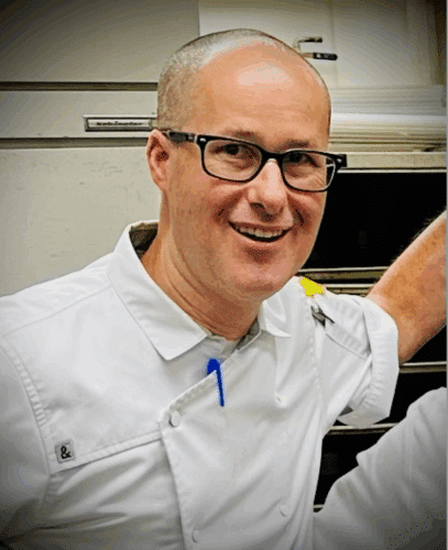 A man wearing glasses and a white chef's coat stands in a kitchen, smiling at the camera. A man wearing glasses and a white chef's coat stands in a kitchen, smiling at the camera.