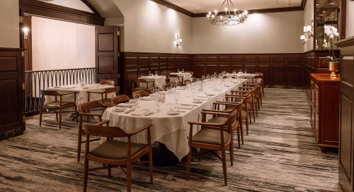 A private dining room with two long tables set for a formal meal, surrounded by wooden chairs, white tablecloths, and wall-mounted lights.